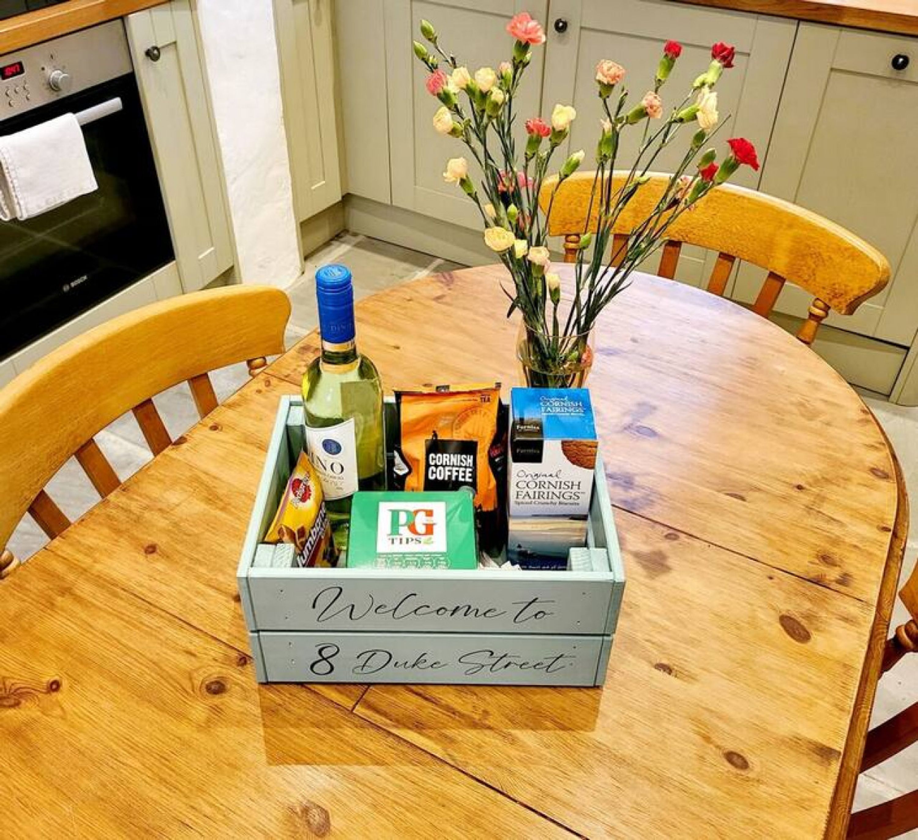 A wooden table with a welcome sign, a bottle of wine, coffee bags, and a box of cornish fairings.