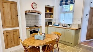 A compact kitchen with a round wooden table and chairs, a built-in oven, a microwave, and a window with a blue and white valance. - 8 Duke Street in Cornwall (Launceston)