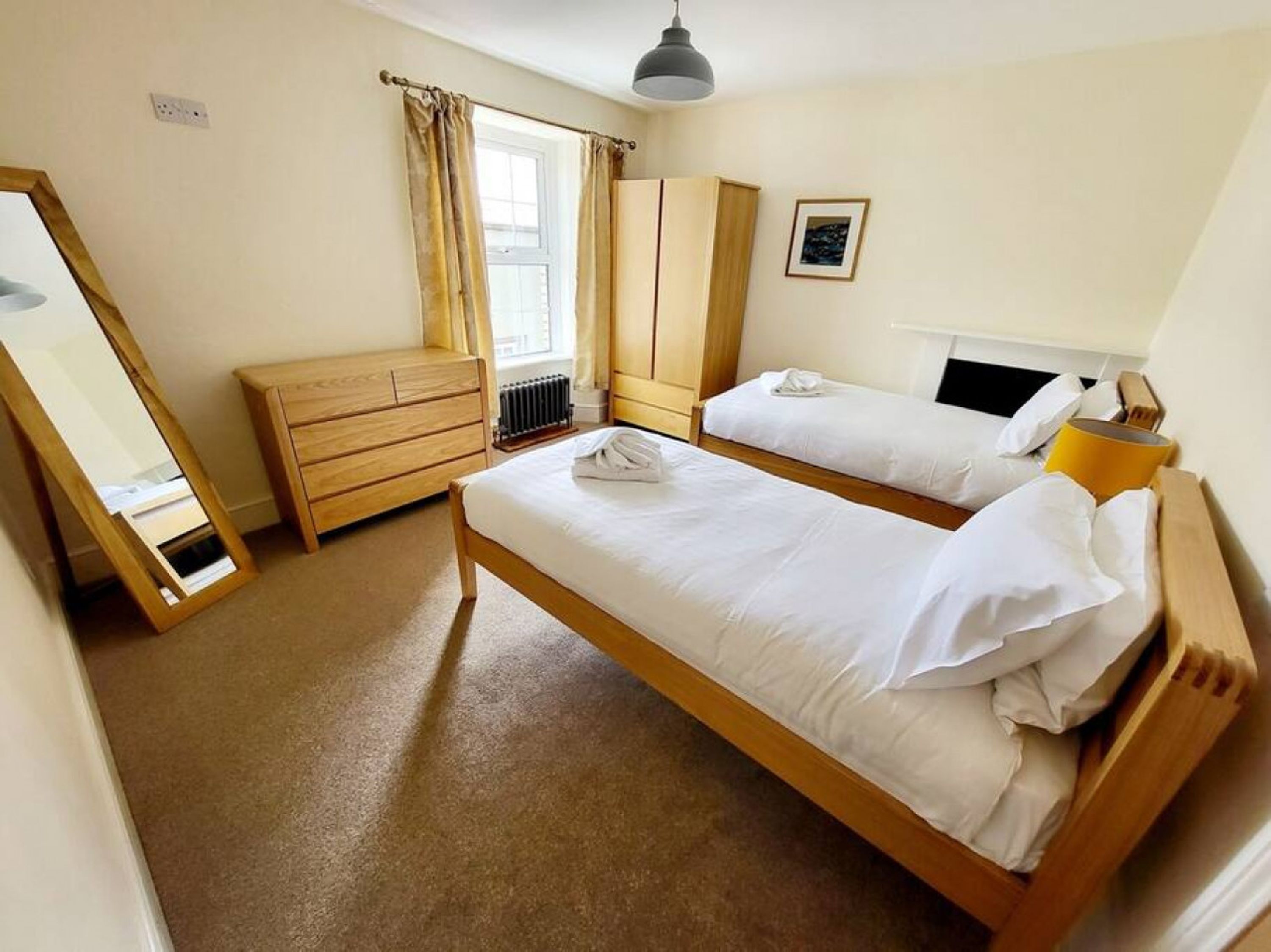 A bedroom with two single beds, a wooden dresser, a mirror, a radiator, and a window with curtains.