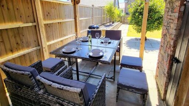 An outdoor patio with a wooden pergola, a table set for dining, wicker furniture, and a brick wall. - 8 Duke Street in Cornwall (Launceston)