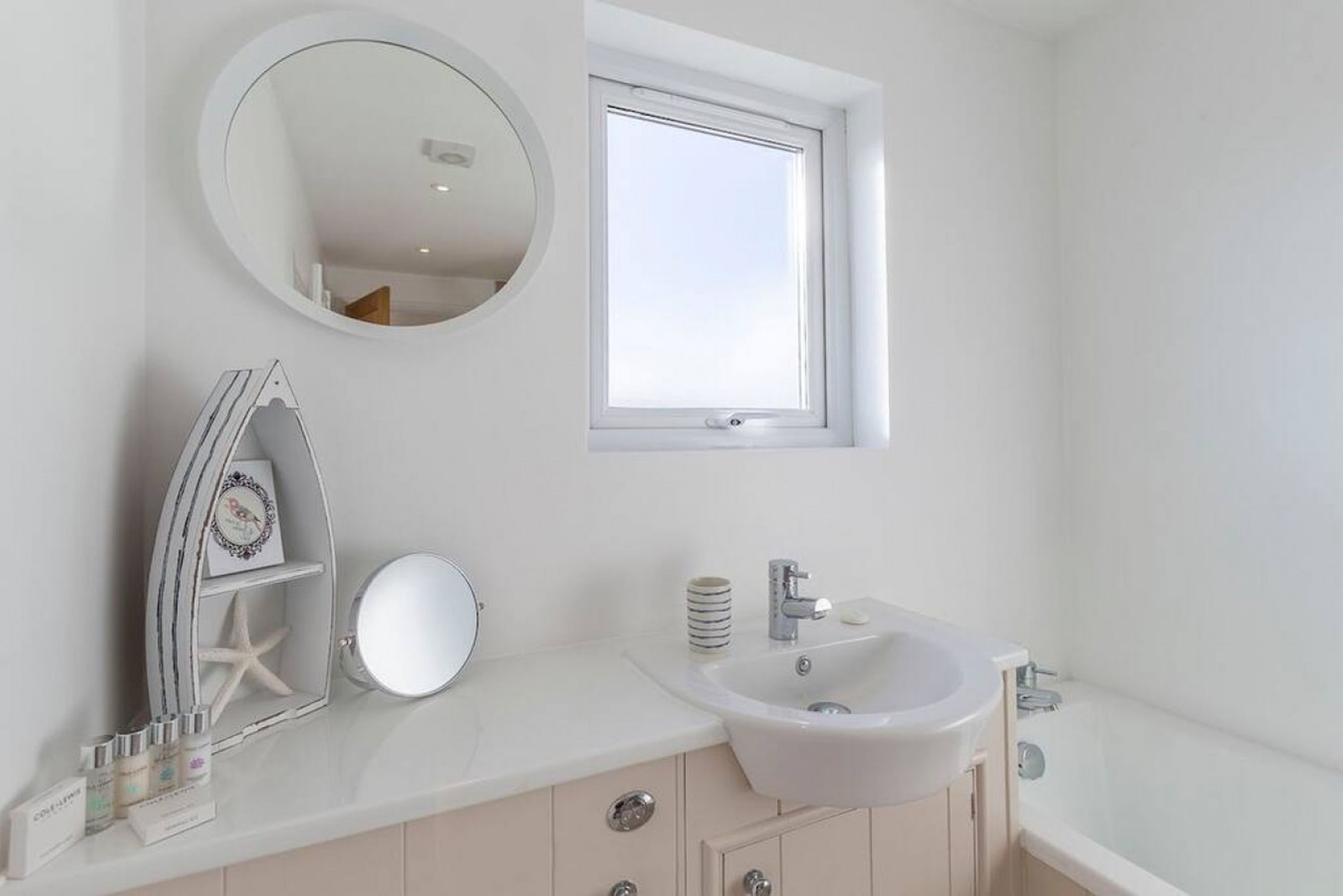 Un baño con un lavabo blanco, un espejo redondo, una decoración de estrella de mar y una ventana.
