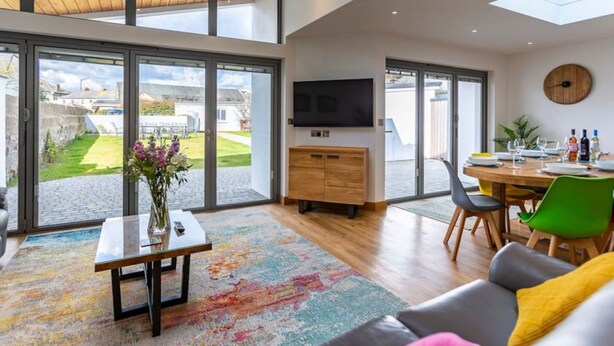A spacious living area with a large window, a flat-screen TV, a dining table with chairs, and a colorful rug. - Number 15 in Cornwall (Bude)