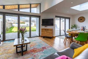 A spacious living area with a large window, a flat-screen TV, a dining table with chairs, and a colorful rug. - Number 15 in Cornwall (Bude)