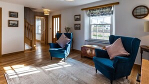 A spacious living room with wooden flooring, two blue armchairs, a wooden coffee table, and a large window with a view. - Number 15 in Cornwall (Bude)