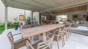 Ein offener Wohnbereich mit einem langen Holztisch und Korbstühlen, einem Sofa und einer Küche im Hintergrund.