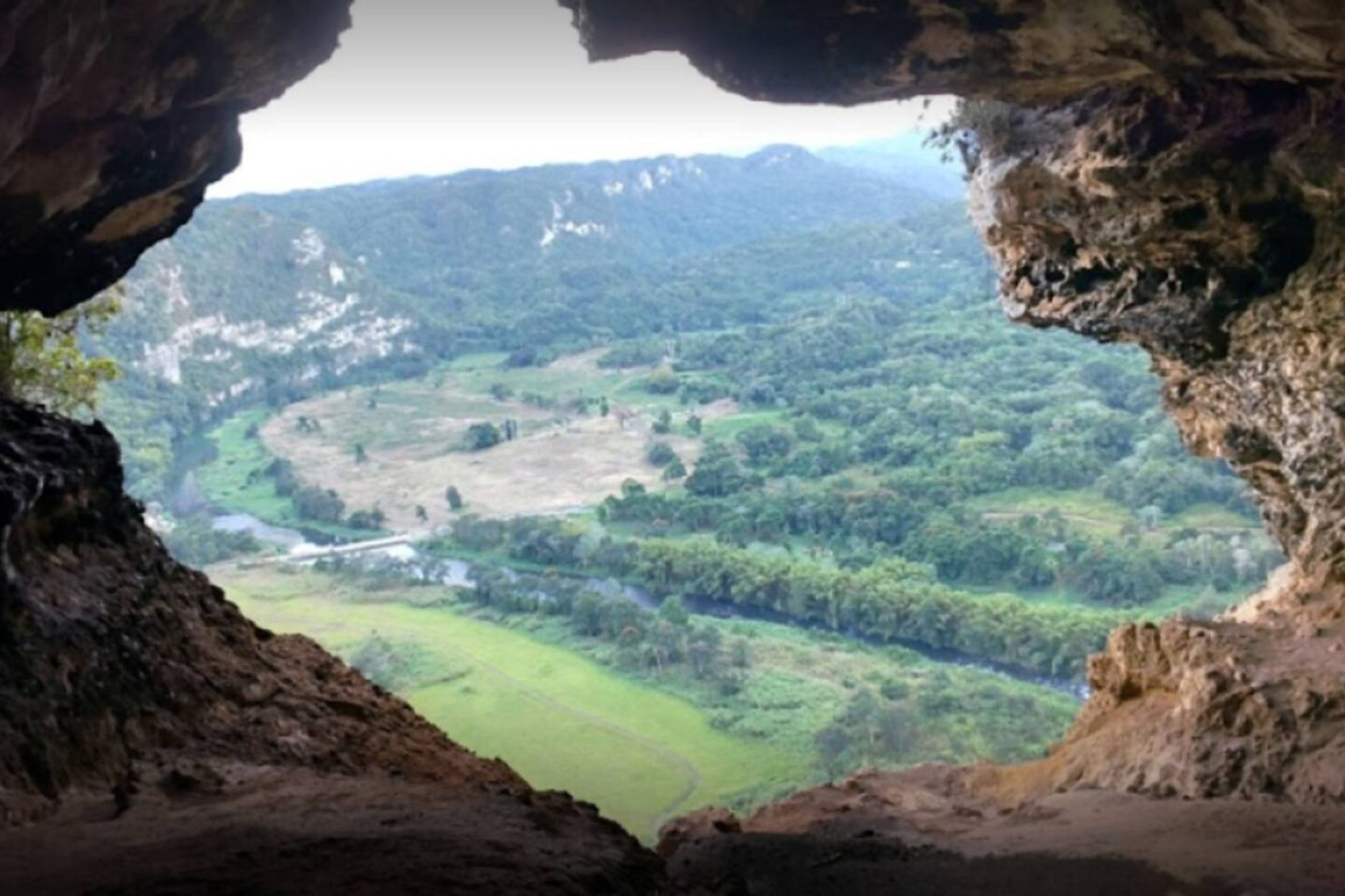 A view through a cave opening, revealing a valley with green fields and distant mountains.