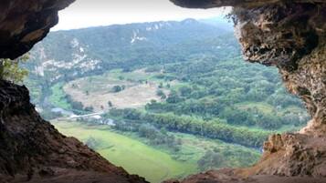 A view through a cave opening, revealing a valley with green fields and distant mountains.