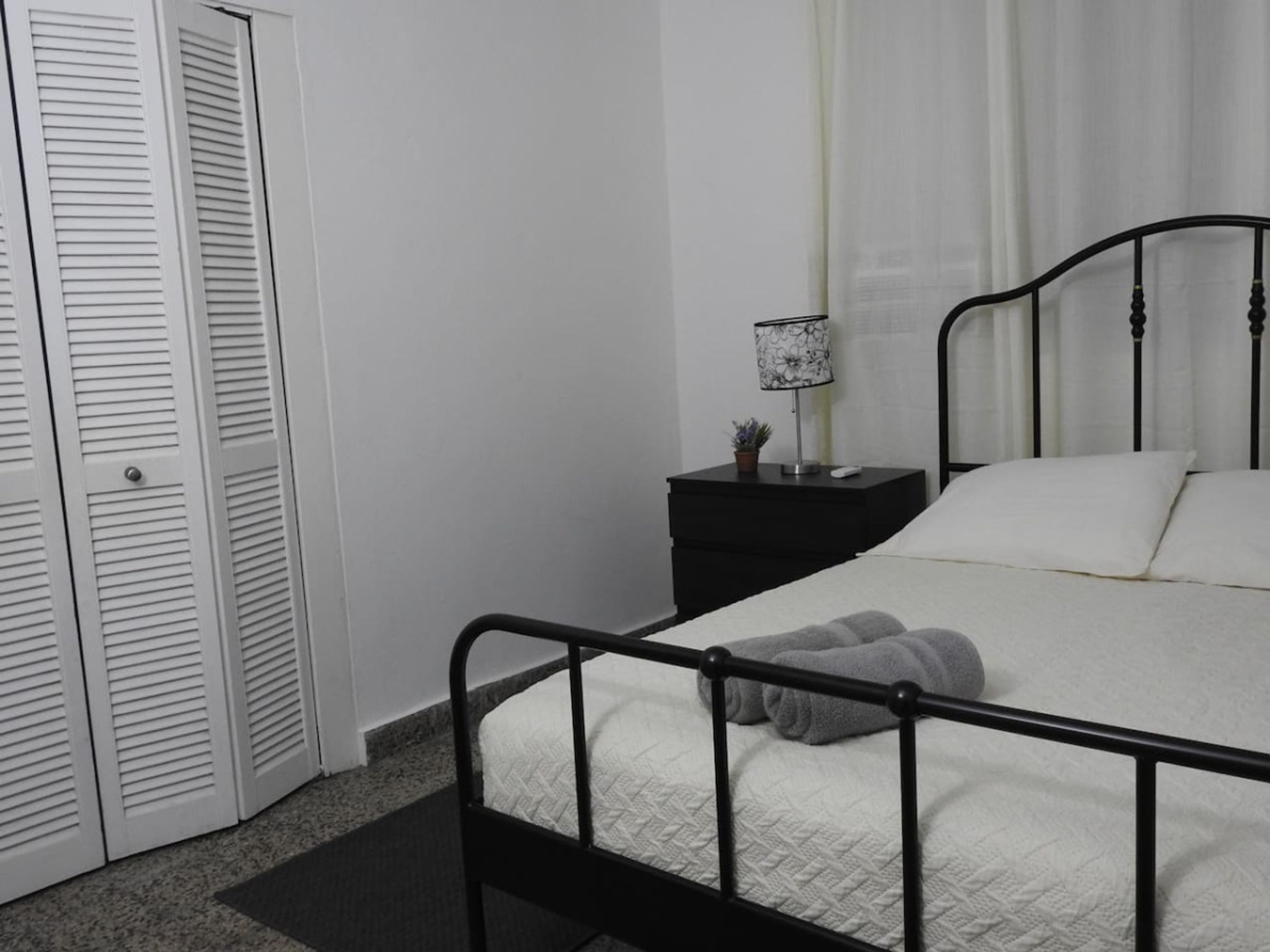 Un dormitorio con estructura de cama metálica, ropa de cama blanca, una mesita de noche con lámpara y un armario blanco cerrado.