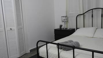 Un dormitorio con estructura de cama metálica, ropa de cama blanca, una mesita de noche con lámpara y un armario blanco cerrado.