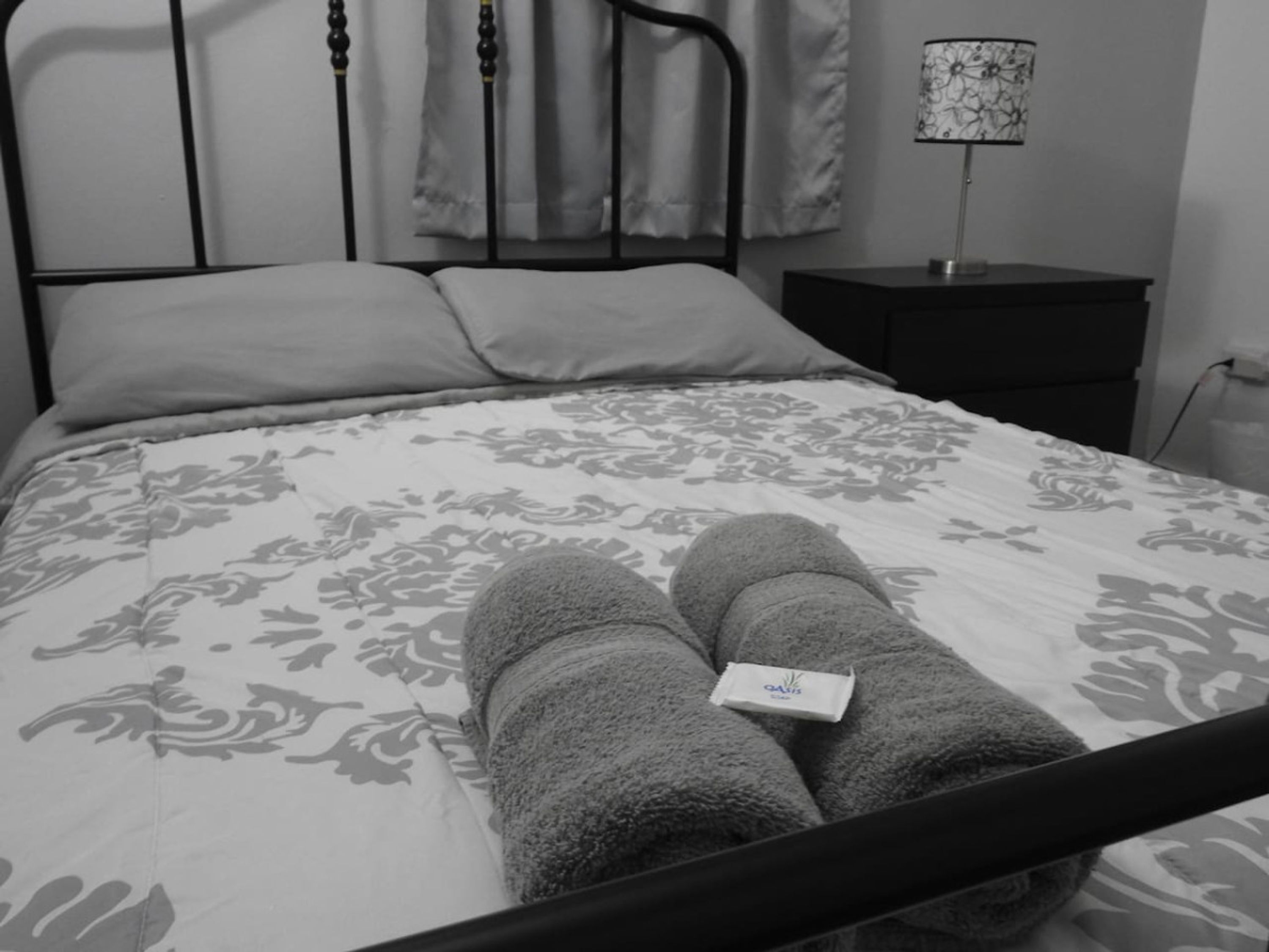 Ein Bett mit floralem Bettbezug, zwei zusammengerollte Handtücher auf dem Bett, eine Nachttischlampe und ein kleiner Nachttisch.