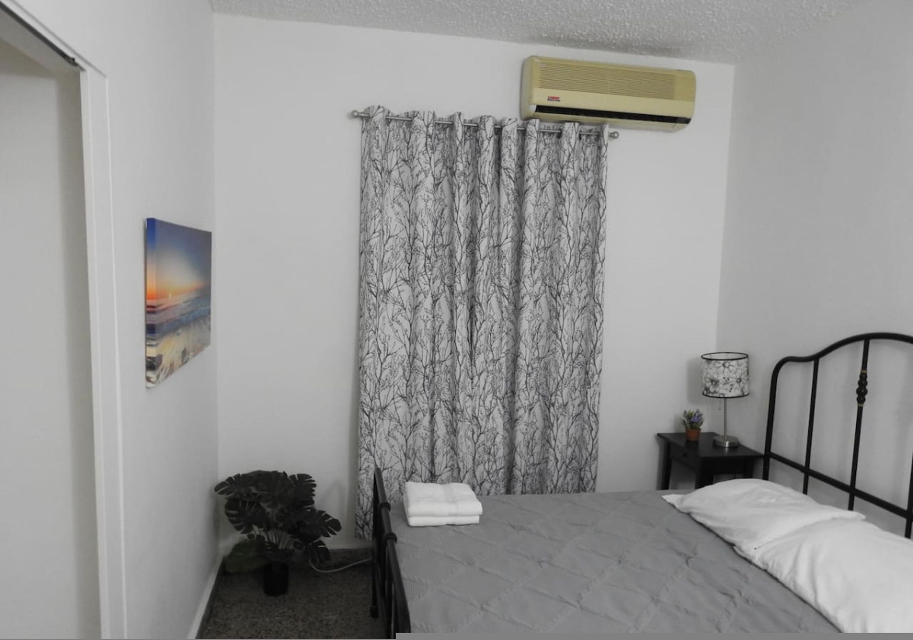 A bedroom with a bed, an air conditioner, a curtain, a nightstand, a lamp, and a painting.