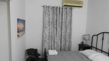 A bedroom with a bed, an air conditioner, a curtain, a nightstand, a lamp, and a painting.