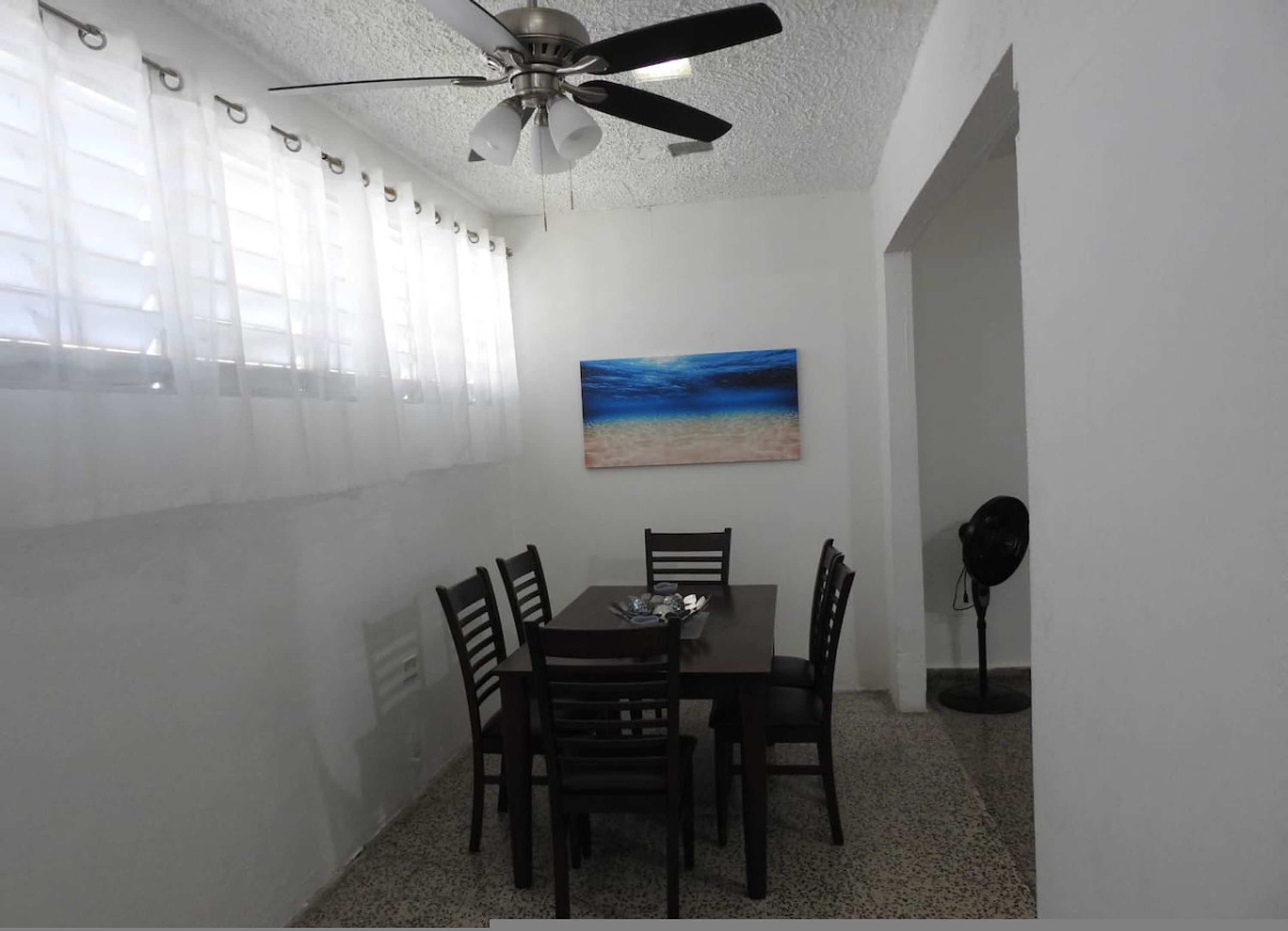 A dining area with a table and chairs, a ceiling fan, a beach painting, and a lamp.