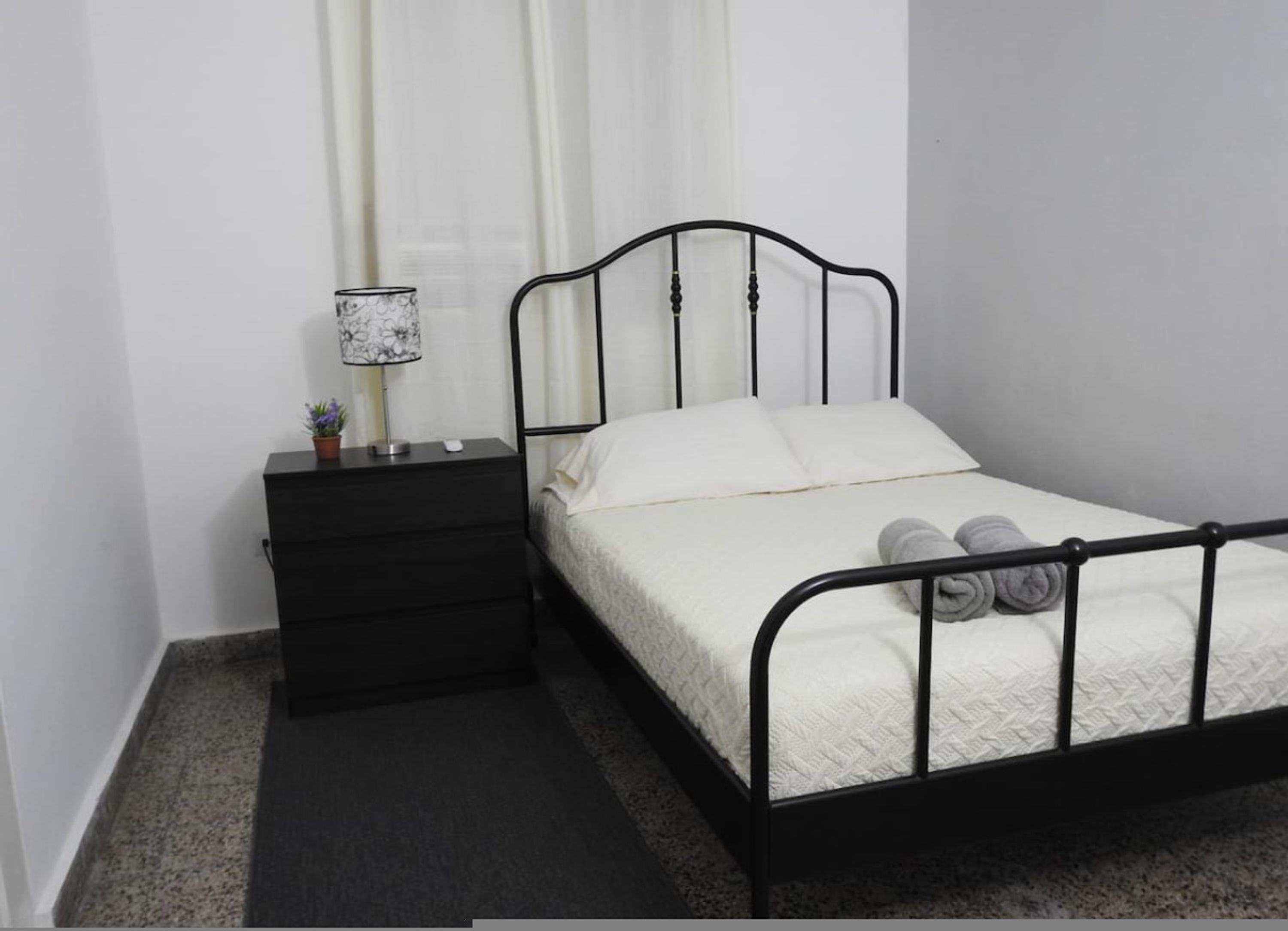 A single bed with a black metal frame, white bedding, and two rolled towels on the footboard.