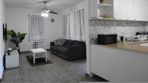 A compact living space with a sofa, a small table, a ceiling fan, a microwave, and a kitchen area with cabinets and a marble backsplash. - Casa del Sol-unit A Island Escape 3BR Near Hospital Malls (Bayamon)