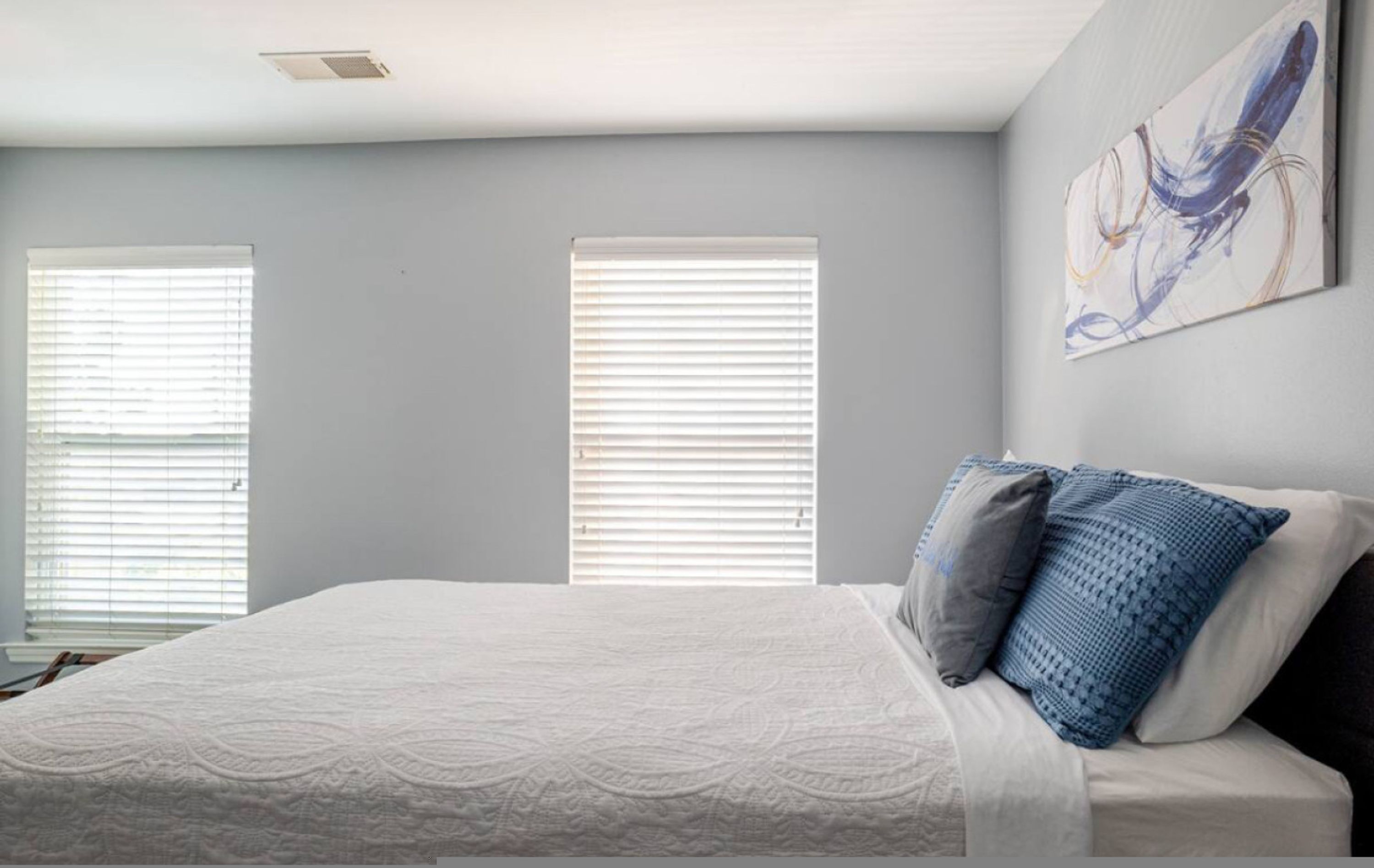 Un dormitorio con una cama grande, dos ventanas con persianas y un cuadro abstracto colgado en la pared.