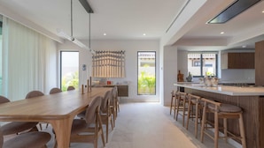 A modern kitchen with a central island, wooden chairs, and a dining area with a long table. - Villa Amistad in Punta Mita (Punta de Mita)