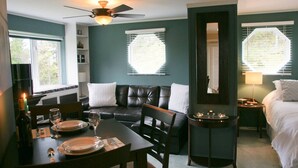 A room with a dining table set for two, a black leather sofa, a bed, and a small side table with a lamp. - Bree s by the Sea - Ocean View Large 2 Person Tub (Nanoose Bay)