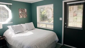 A bedroom with a bed, a nightstand, a window with blinds, a door with a glass panel, and a wall with a decorative piece. - Bree s by the Sea - Ocean View Large 2 Person Tub (Nanoose Bay)