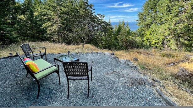 An outdoor seating area with two chairs and a table, surrounded by trees and a clear sky. - Bree s by the Sea - Ocean View Large 2 Person Tub (Nanoose Bay)