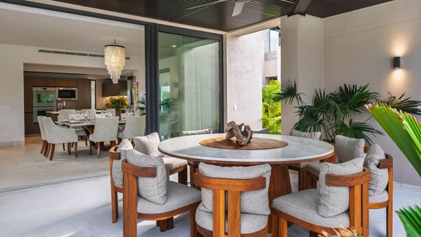 A modern dining area with a round wooden table and cushioned chairs, a chandelier, and a view of the kitchen and living spaces. - Villa Amada in Punta Mita (Punta de Mita)