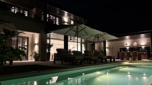 A modern outdoor pool area with lounge chairs and umbrellas, a dining area with a kitchen, and a large house with multiple windows and a balcony. - Villa Amada in Punta Mita (Punta de Mita)