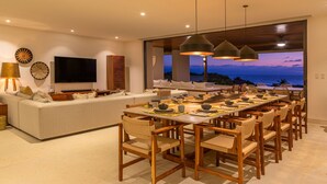A modern living room with a large TV, a dining area with wooden chairs and a table, and a view of the ocean at dusk. - Condo Haramara in Punta Mita (Punta de Mita)