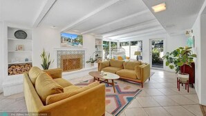 A spacious living room with a fireplace, yellow sofas, and a large window offering a view of the outdoors. - Castaways Casita Saltwater Pool Dog Friendly (Fort Lauderdale)