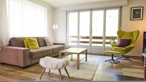Sala de estar com sofá, uma mesinha, um banquinho macio, uma poltrona verde e vista através de uma porta de correr.