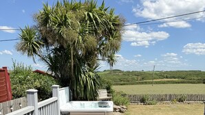 A wooden deck with a hot tub, a wicker sofa, and a view of a field and hills. - Meadow View in Widemouth Bay (Bude)