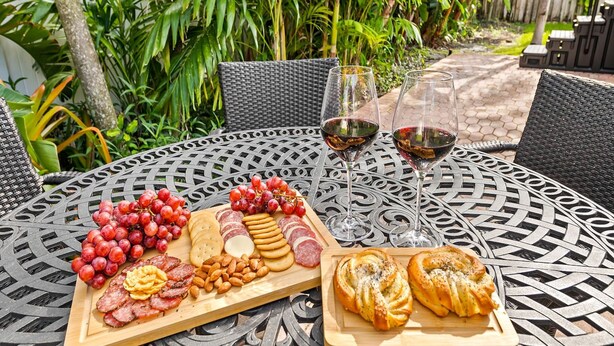 An outdoor dining area with a metal table, a charcuterie board, and two glasses of red wine. - Beach Bungalow Walk to Beach Swimspa Jacuzzi (Lauderdale-by-the-Sea)