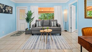 A living room with a grey sofa, two small round tables, a ceiling fan, and a view of greenery outside. - Beach Bungalow Walk to Beach Swimspa Jacuzzi (Lauderdale-by-the-Sea)