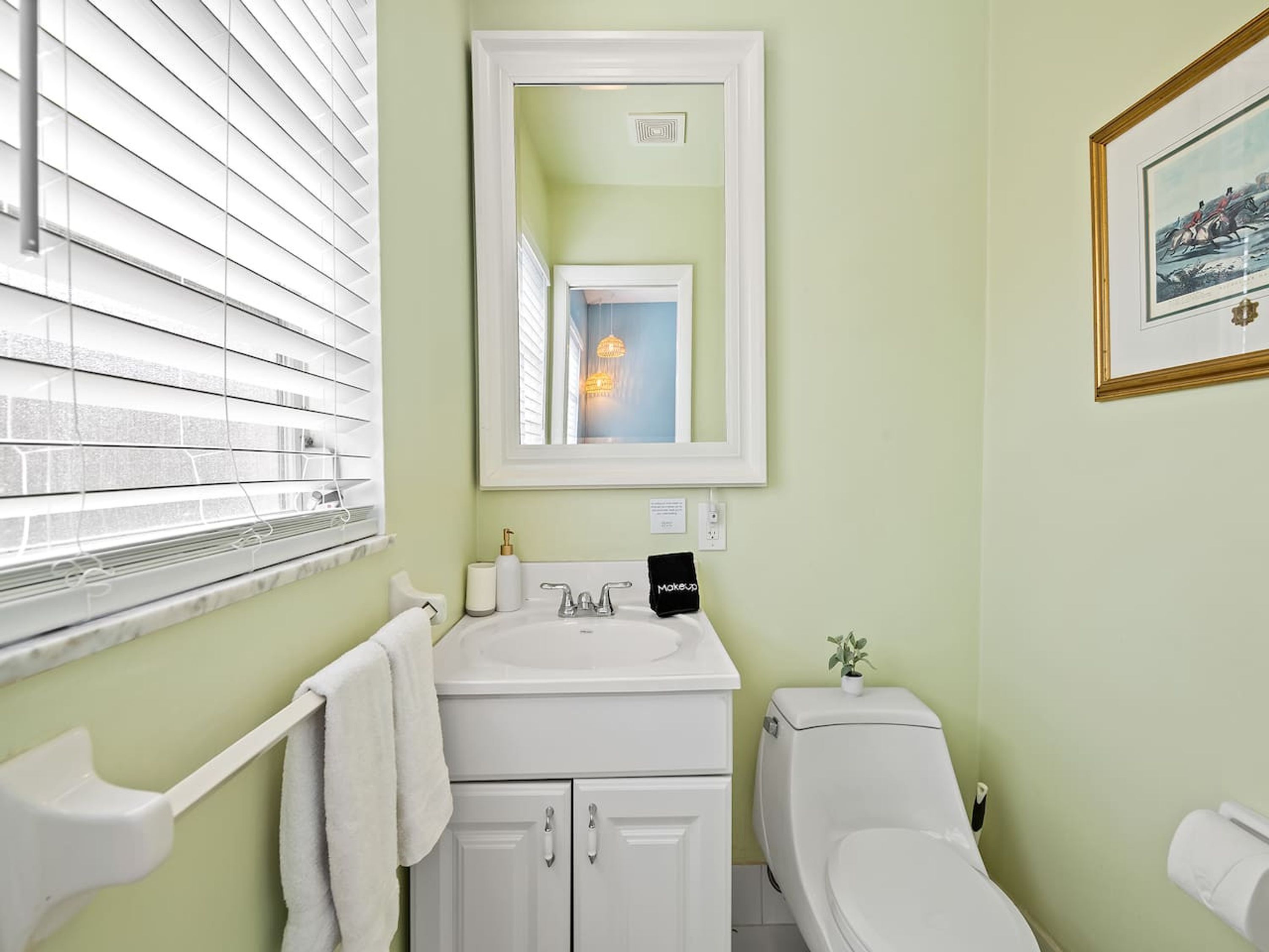 A bathroom with a white sink, a mirror above it, a toilet, and a window with blinds.