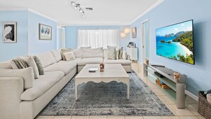 A living room with a beige sectional sofa, a white coffee table, a blue accent wall, a ceiling fan, and a TV displaying a scenic view. - Beach Bungalow Walk to Beach Swimspa Jacuzzi (Lauderdale-by-the-Sea)