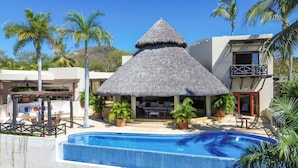 A resort with a swimming pool, a thatched-roof structure, and palm trees. - Casa Caracol - Mita Residential Farallon (Punta de Mita)