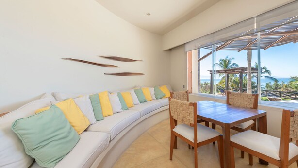 A modern living room with a curved white sofa, a wooden dining table with chairs, and a large window offering a view of the ocean and palm trees. - Casa Caracol - Mita Residential Farallon (Punta de Mita)