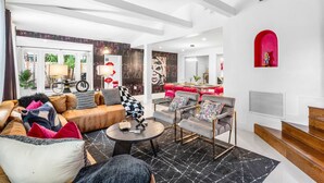 A modern living room with a sofa, armchairs, and a coffee table. There is a dining area with a red table and chairs. The room has white walls and a red archway. - Chic Retreat Heated Pool Floats Close to Beach (Fort Lauderdale)