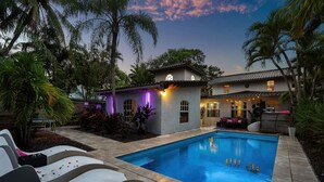 A swimming pool with lounge chairs, a small building with a lighted exterior, and palm trees. - Chic Retreat Heated Pool Floats Close to Beach (Fort Lauderdale)