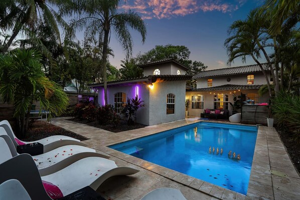 A swimming pool with lounge chairs, a small building with a lighted exterior, and palm trees.