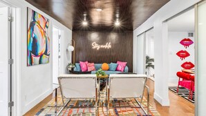 A modern living room with a wooden ceiling, a colorful abstract painting, a sofa with pink and blue cushions, and a neon sign that reads "Stay awhile". - Chic Retreat Heated Pool Floats Close to Beach (Fort Lauderdale)
