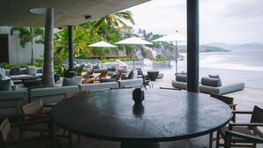 A modern outdoor seating area with a round table, lounge chairs, and umbrellas, overlooking a beach with palm trees and a rocky cliff. - Valhalla Luxury Villa (Punta de Mita)