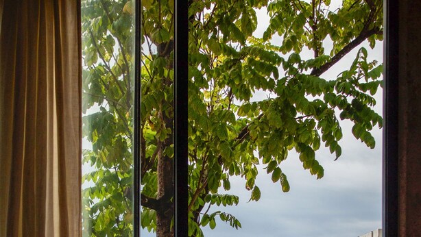 A round table with a single chair, a window with curtains, and a view of a tree and water. - Valhalla Luxury Villa (Punta de Mita)