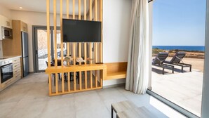 A modern living room with a TV mounted on a wooden partition, a dining area, and a large window offering a view of the sea. - Isanema Villa 2 in Karpathos (Karpathos)