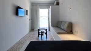 A modern hotel room with a bed, a small round table, a sofa, a TV mounted on the wall, and a large window with a view. - Aerase Apartment in Karpathos (Karpathos)