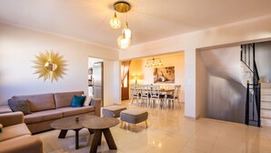 A modern living room with a sofa, coffee table, and ottoman. There is a dining area with a table and chairs. A kitchen is visible in the background. - Villa Danai in Volada (Karpathos)