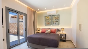 A bedroom with a large bed, two bedside tables, a sliding glass door leading to an outdoor area, and two framed artworks on the wall. - Isanema Villa 1 in Karpathos (Karpathos)