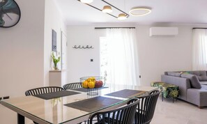 A modern living room with a glass dining table, black chairs, a grey sofa, and a wall-mounted air conditioner.