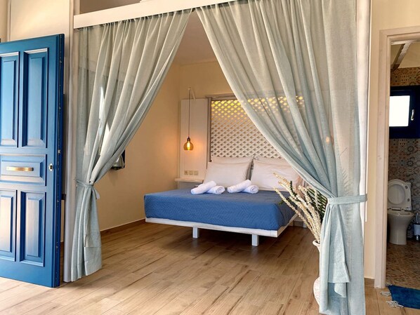 A bedroom with a bed, curtains, a bedside lamp, and a bathroom visible through an open door. - Michail Suites Afiartis Vatha (Karpathos)
