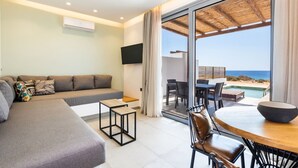 A modern living room with a sofa, coffee table, and dining area. A TV is mounted on the wall, and there is a large sliding glass door leading to an outdoor patio with a view of the ocean.