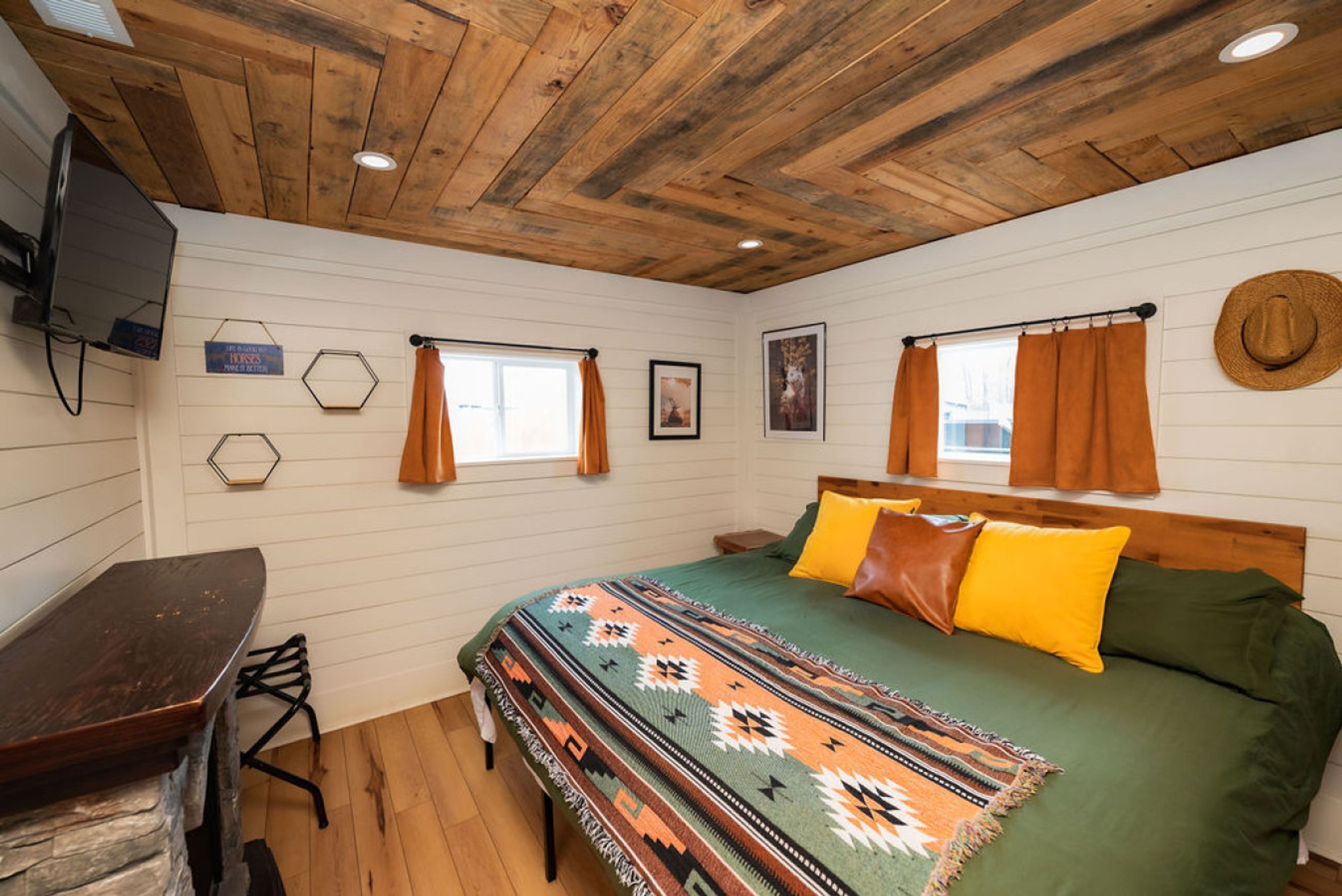 A bedroom with a wooden ceiling, a bed with a patterned blanket, a desk, and a window with curtains.